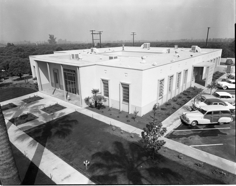 Historic photo of the single story building for the Redlands Daily Facts, with cars in a parking lot on the left and surrounded by orange groves. 