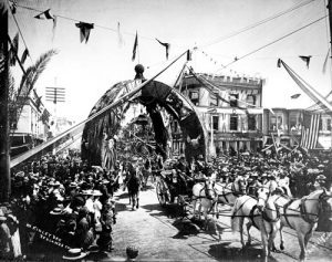 A photograph of the intersection of State and Orange Streets decorated for President William McKinley's visit.  A large arch is in the center of the intersection, with a horse-drawn carriage in the street, hundreds of spectators along the streets, and flags and bunting on business buildings. 