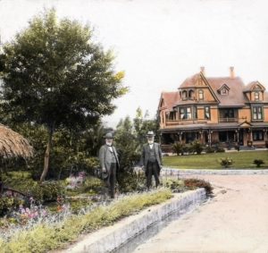 Colorized photo of two identical twins with white beards in suits and hats among plants in front of a large Victorian house.