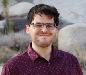 Portrait of a man with brown hair and glasses wearing a maroon shirt.  